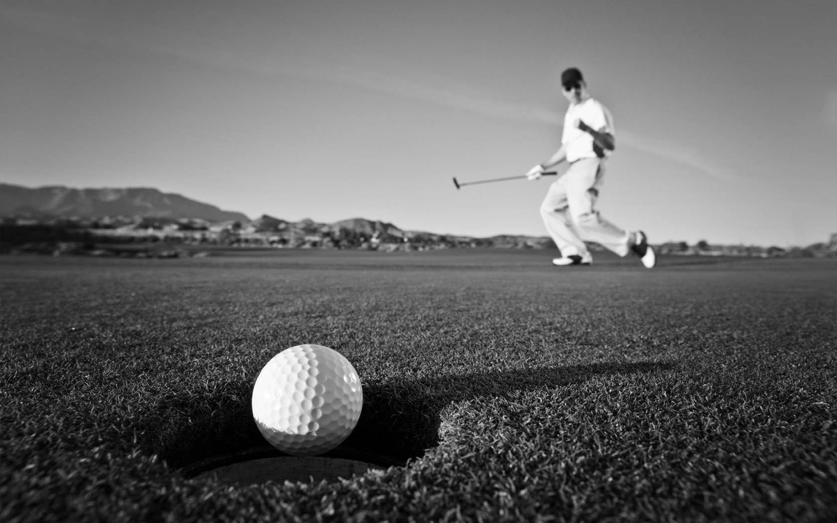 Schwarz-Weiß-Foto: ein Golfball am Rand des Lochs, dahinter ein Golfer